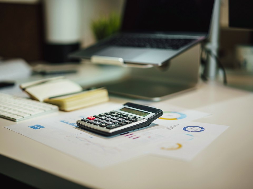 A calculator resting on printed financial charts on a desk, with a laptop and notebook behind it