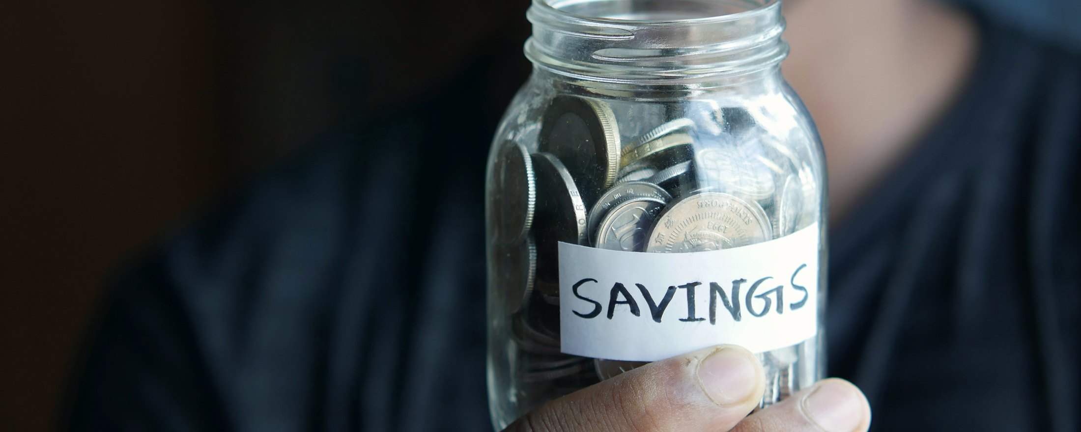 A hand holding a glass jar of coins labeled Savings